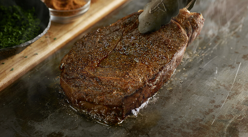 Picture of a steak being cooked on a skillet with ingredients in the background