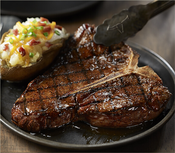 Steak being laid onto a plate by tongs with a loaded baked potato