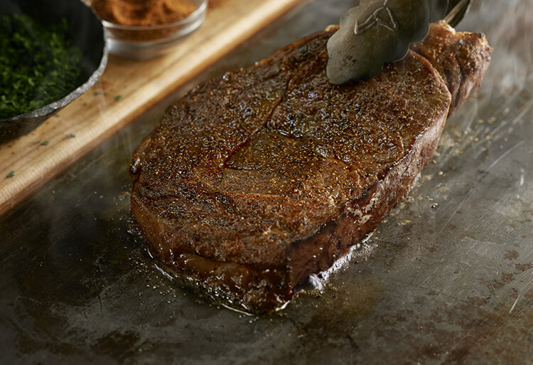 Picture of a steak being cooked on a skillet with ingredients in the background