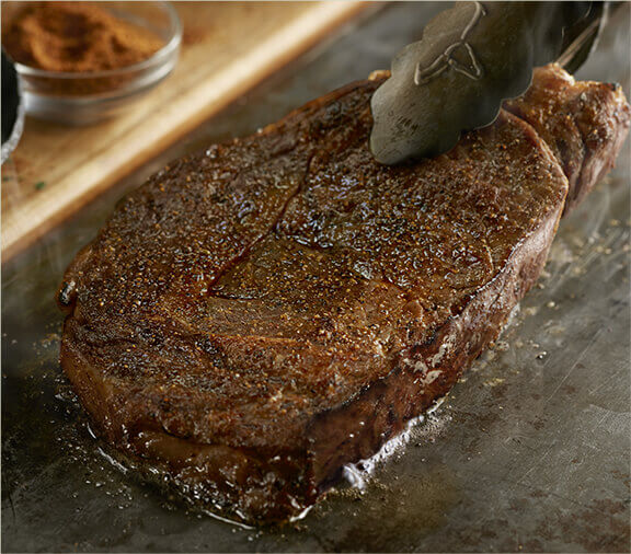 Boneless ribeye being pressed on a grill by tongs